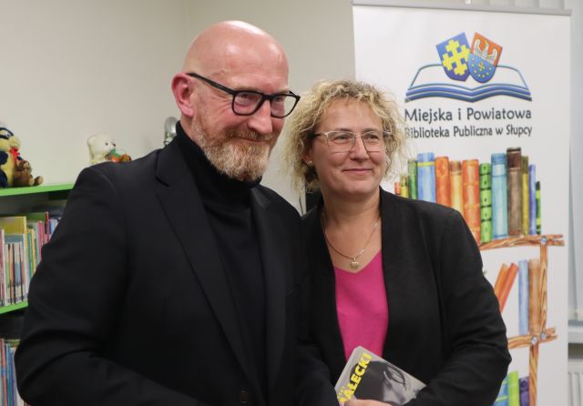 Na zdjęciu znajduje się kobieta stojąca do zdjęcia z mężczyzną. W tle znajdują się półki z książkami oraz baner z logiem biblioteki i książkami. 