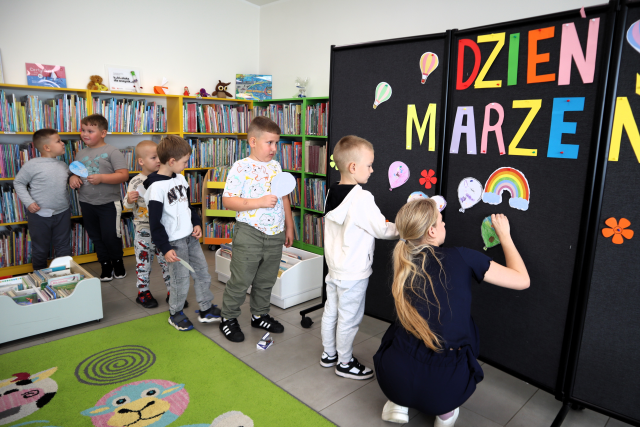 Na zdjęciu znajdują się dzieci, które stoją jeden za drugim i przyklejają do czarnej tablicy z napisem swoje balony. W tle znajdują się półki z książkami.