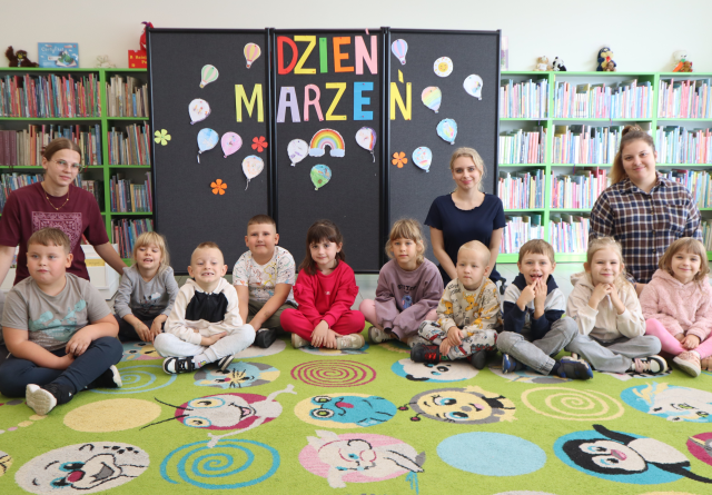 Na zdjęciu znajduje się 10 przedszkolaków oraz trzy kobiety i siedzą razem na dywanie do wspólnego zdjęcia, za nimi znajduje się tablica z napisem i pracami wykonanymi na zajęciach. W tle znajdują się półki z książkami.