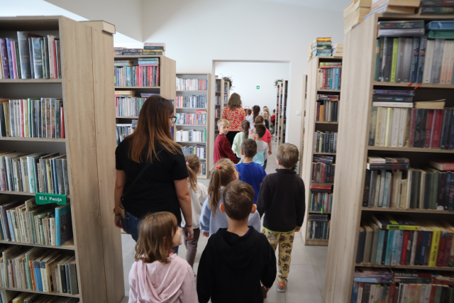 Na zdjęciu znajduje się grupa przedszkolna praz trzy kobiety, w wypożyczalni bibliotecznej i idą między półkami z książkami.