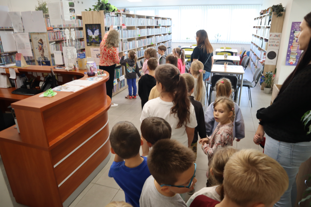 Na zdjęciu znajdują się przedszkolaki oraz trzy kobiety, jedna z kobiet pokazuje dzieciom działy z książkami na półkach, po lewej stronie znajduje się biurko.