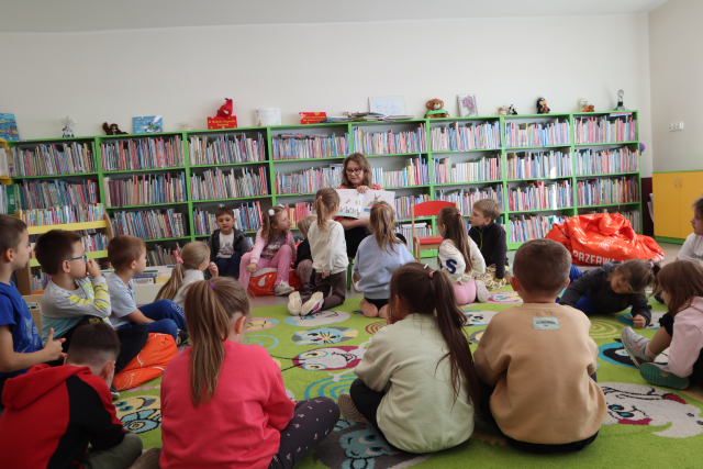 Na zdjęciu znajdują się dzieci na dywanie, a przed nimi siedzi kobieta i pokazuje im ilustracje z książki. W tle znajdują się półki z książkami.