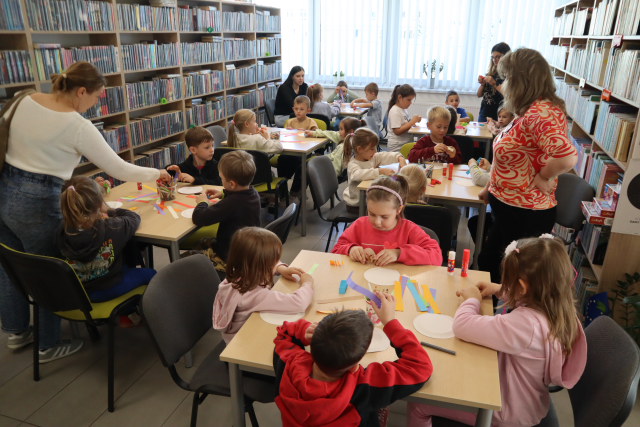 Na zdjęciu znajdują się dzieci przy stolikach, przy których wykonują prace plastyczną, przy stolikach stoją trzy kobiety. W tle znajdują się półki z książkami oraz okna.