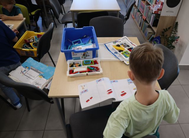 Na zdjęciu znajduje się chłopiec przy stoliku przed sobą ma instrukcje oraz klocki LEGO w pudełku. Na przeciwko oraz po lewej stronie znajdują się stoliki z krzesełka, na jednym z nich po lewej stronie leży pudełko z klockami oraz przy stoliku siedzi dwóch chłopców.