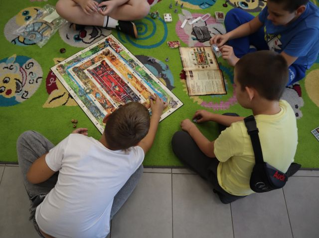 Na zdjęciu znajduje się czterech chłopców na dywanie i grają w grę planszową, przed sobą mają planszę, karty oraz instrukcję.