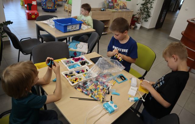 Na zdjęciu znajduje się czterech chłopców, przy stoliku w centrum siedzi 3 chłopców i układają wspólnie klocki LEGO, przy drugim stoliku siedzi chłopiec, który układa klocki sam. W tle znajduje się szafka, kwiaty.
