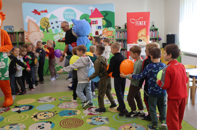 Na zdjęciu znajdują się dwa pociągi tworzone przez przedszkolaków oraz maskotki lisa oraz niebieskiego kota, pomiędzy sobą na poziomie brzucha trzymają balon. W tle znajdują się półki z książkami oraz baner z postaciami z książki na placu zabaw, obok stoi czerwony baner z postacią lisa.