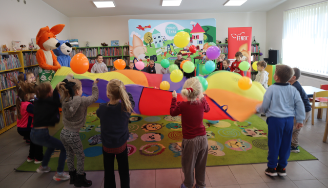 Na zdjęciu znajdują się przedszkolaki trzymające kolorową chustę wraz z maskotką lisa i niebieskiego kota i podrzucają znajdujące się tam balony. W tle znajduje się baner z postaciami z książki, obok czerwony baner z postacią lisa oraz półki z książkami.