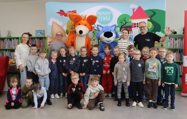 Na zdjęciu znajdują się przedszkolaki wraz z 2 paniami, panem oraz maskotką lisa i niebieskiego kota, wszyscy stoją do wspólnego zdjęcia na tle baneru przedstawiającego postacie z książki na placu, w tle znajdują się półki z książkami.