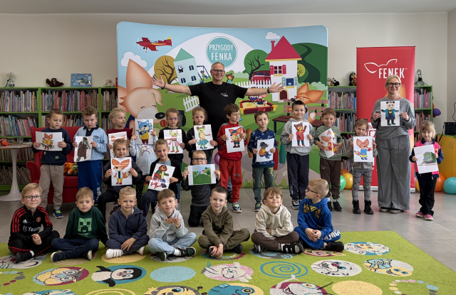 Na zdjęciu znajdują się przedszkolaki, część z nich kuca, część stoi wraz z mężczyzną i kobietą. Część dzieci trzyma w ręce kartkę z bohaterami książek. Za nimi znajduje się baner przedstawiający bohaterów książkowych na placu zabaw, po prawej stronie stoi czerwony baner z lisem. W tle znajdują się półki z książkami.