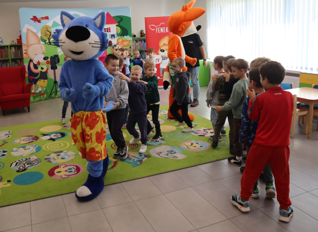 Na zdjęciu znajdują się przedszkolaki, które robią pociąg za maskotką lisa i niebieskiego kota. W tle znajdują się półki z książkami, czerwony fotel, baner z postaciami z książek oraz czerwony baner z postacią lisa.