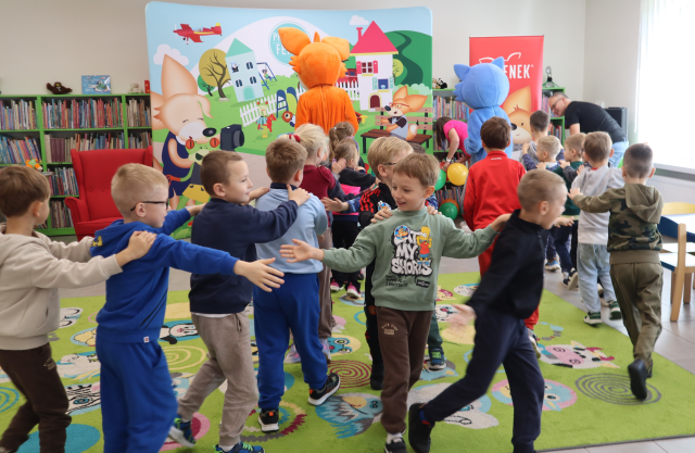 Na zdjęciu znajdują się przedszkolaki, które robią pociąg w dwóch rzędach i przybijają sobie piątki, pociągi prowadzą dwie maskotki lis i niebieski kot. W tle znajdują się półki z książkami, fotel oraz baner z postaciami z książki na placu zabaw.