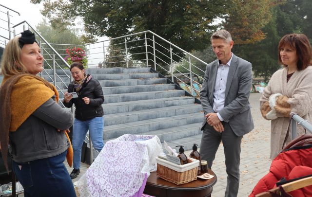 Na zdjęciu znajduje się kobieta i mężczyzna po prawej stronie i oglądają rzeczy wystawione przez kobietę stojącą po lewej stronie. W tle znajduje się kobieta robiąca zdjęcie telefonem, schody oraz drzewa.