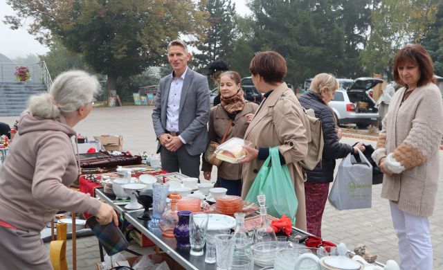 Na zdjęciu znajduje się mężczyzna oraz 4 kobiety, trzy z nich oglądają rzeczy wystawione przez kobietę po lewo, naczynia, biżuterię. W tle znajduje się parking oraz drzewa.