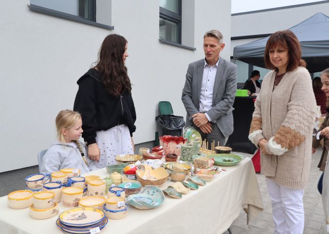 Na zdjęciu znajduje się mężczyzna oraz kobieta, którzy stoją przy stoiska dziewczynki oraz nastolatki, które na stoliku przed sobą mają ceramikę. W tle znajduje się budynek.