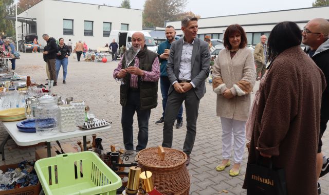 Na zdjęciu znajduje się grupka ludzi oglądających wystawione rzeczy na sprzedasz, w tym koszyki, wazony, jeden z mężczyzn trzyma duży przezroczysty wazon. W tle znajduje się parking z wystawionymi sprzedawcami oraz odwiedzającymi oraz budynek.