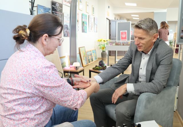 Na zdjęciu znajduje się kobieta robiąca masaż dłoni mężczyźnie, który siedzi naprzeciwko niej na fotelu. W tle znajdują się stoliki z olejkami, obrazy, plakaty na ścianach oraz słoneczniki w wazonie.