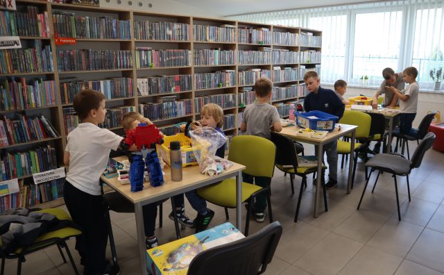 Na zdjęciu znajduje się 9 chłopców przy trzech stolikach i układają klocki LEGO znajdujące się w pudełkach na stolikach. W tle znajdują się półki z książkami oraz okna.