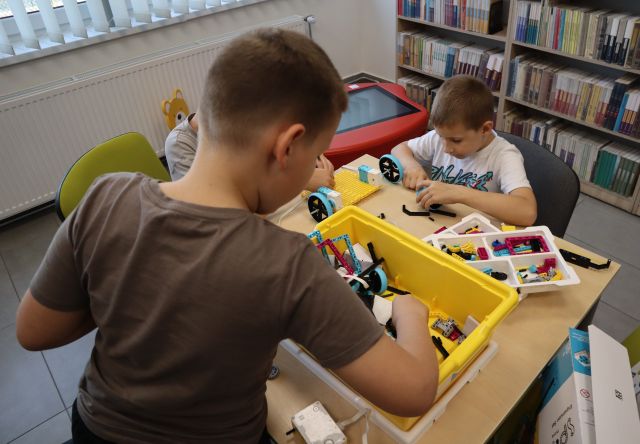 Na zdjęciu znajduje się 3 chłopców przy stoliku i układają współnie klocki LEGO, jeden z chłopców stoi i sięga potrzebne elementy z pudełka. W tle znajdują się półki z książkami i czerwony stolik interaktywny.