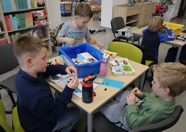 Na zdjęciu znajduje się trójka chłopców przy stoliku i układają klocki LEGO według instrukcji, na stoliku leży również niebieskie pudełko na klocki. W tle znajdują się półki z książkami oraz stolik po prawej stronie przy którym siedzi chłopiec i układa klocki.