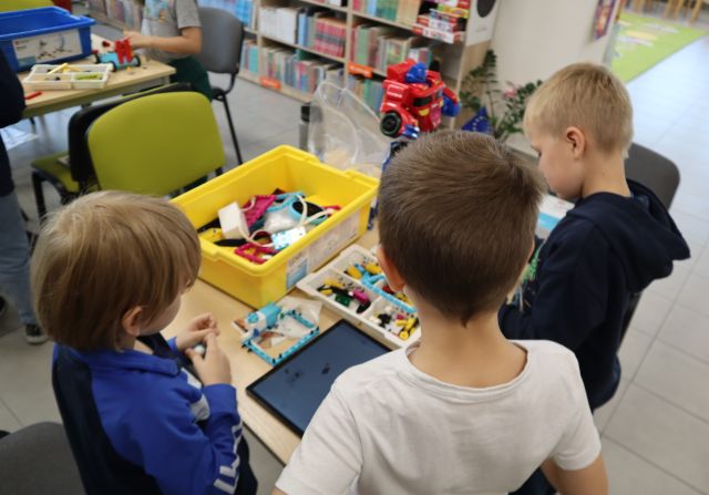 Na zdjęciu znajduje się trójka chłopców przy stoliku i układają wspólnie klocki LEGO według instrukcji na tablecie, na stoliku przed nimi stoi żółte pudełko z klockami LEGO. W tle znajdują się półki z książkami.