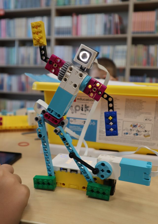 Na zdjęciu znajduje się robot zbudowany z klocków LEGO, który tańczy, za nim znajduje się pudełko z klockami oraz klocki na stoliku. W tle znajdują się półki z książkami.