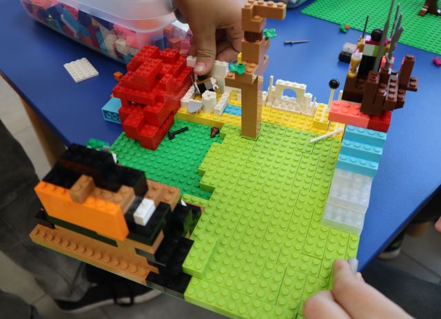 Na zdjęciu znajduje się konstrukcja z klocków LEGO przedstawiająca las, fortecę. Praca znajduje się na niebieskim stoliku.