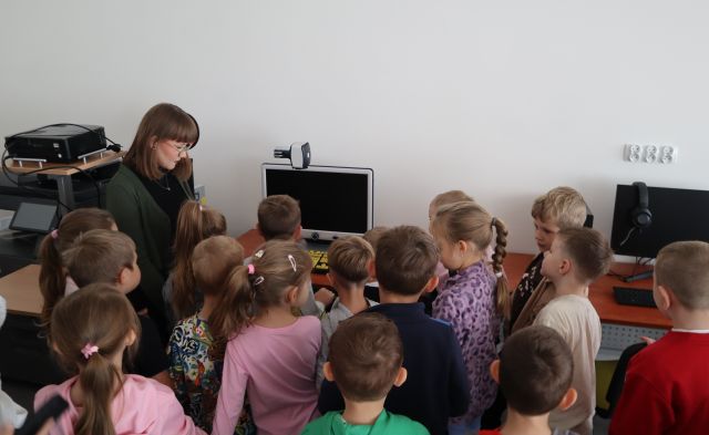 Na zdjęciu znajdują się przedszkolaki w grupie, przed nimi stoi kobieta i pokazuje komputer. Grupa znajduje się w sali komputerowej.