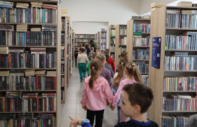 Na zdjęciu znajduje się grupa, wraz z kobieta między regałami z książkami w bibliotece.