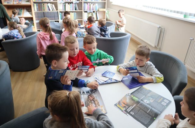 Na zdjęciu znajdują się przedszkolaki przy stolikach w czytelni i przeglądają magazyny. W tle znajdują się półki z książkami.
