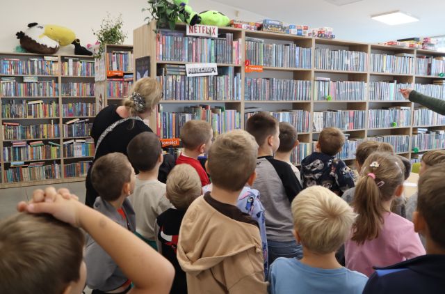Na zdjęciu znajdują się przedszkolaki oraz dwie kobiety w sali bibliotecznej, jedna kobiet stojąca przed nimi pokazuje im ręką półkę. W tle znajdują się półki z książkami.
