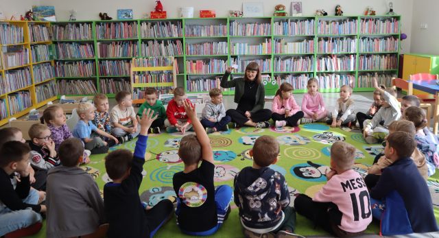Na zdjęciu znajdują się przedszkolaki wraz z kobietą i siedzą na dywanie w okręgu, kobieta pokazuje dzieciom kartę biblioteczną, a część dzieci podnosi rękę. W tle znajdują się półki z książkami.