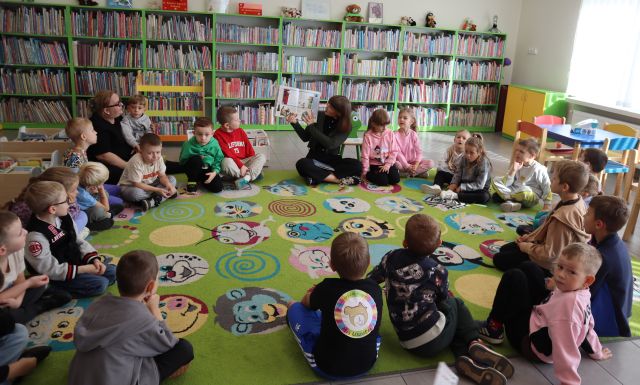 Na zdjęciu znajduje się 24 przedszkolaków siedzących w okręgu na dywanie wraz z kobietą, która pokazuje dzieciom ilustracje z książki. W tle znajdują się półki z książkami.