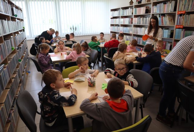 Na zdjęciu znajduje się 24 przedszkolaków przy 6 stolikach, pomiędzy stolikami stoją trzy kobiety. Dzieci przy stolikach robią koperty z kolorowych kartek. W tle znajdują się półki z książkami.