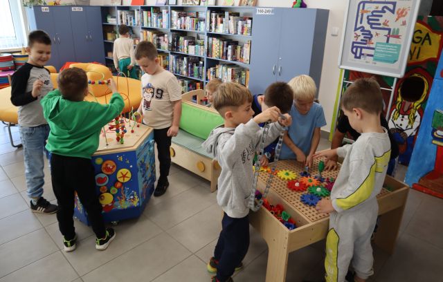 Na zdjęciu znajdują się przedszkolaki w sali bibliotecznej, które się bawią stolikiem do majsterkowania, samochodzikiem oraz stolikiem kreatywnym, w tle znajdują się półki z grami.