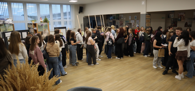 Na zdjęciu znajduje się kolejka osób czekających na spotkanie z autorką na holu biblioteki. W tle znajdują się okna, obrazy ora plakaty na ścianach.