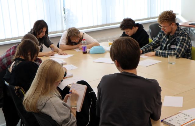 Na zdjęciu znajduje się 8 młodzieży przy złączonych stołach w sali komputerowej, mają przed sobą kartki, a część z nich na nich pisze. W tle znajdują się okna.