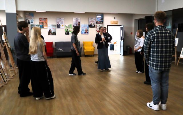 Na zdjęciu znajduje się młodzież na holu biblioteki wraz z kobietą, któa stoi po środku. Po lewej i prawej stronie znajdują się sztalugi z obrazami. W tle znajduje się kanapa z fotelami, plakaty na ścianach oraz drzwi.