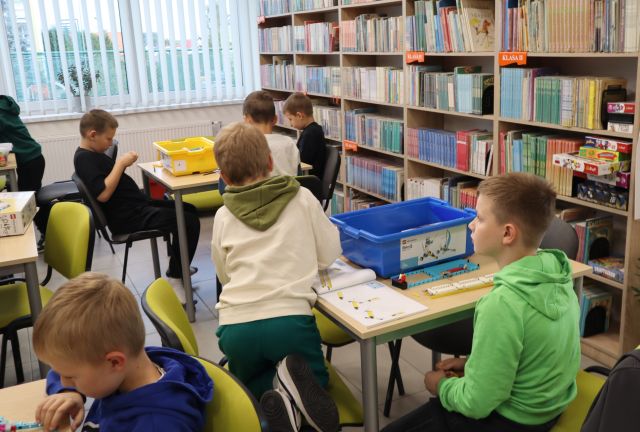 Na zdjęciu znajduje się 5 chłopców przy stolikach przy których układają klocki LEGO, po prawej stronie znajdują się półki z książkami, a po lewej trzy stoliki, przy pierwszym i ostatnim siedzi chłopiec.