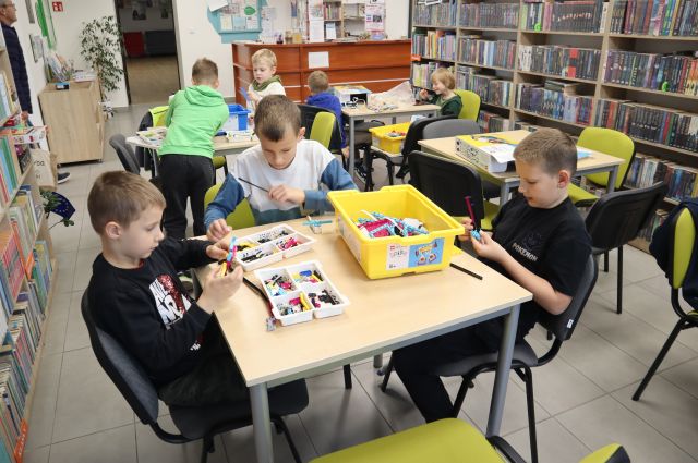 Na zdjęciu znajduje się 7 chłopców przy trzech stolikach, czwarty stolik stoi pusty po prawej stronie. Chłopcy układają klocki lego znajdujące się w pudełkach. W tle znajdują się półki z książkami i biurko.