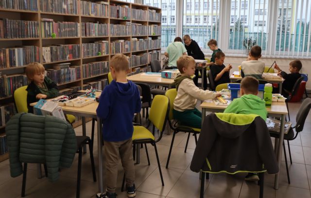 Na zdjęciu znajduje się 5 stolików, przy 4 siedzi 11 chłopców i układają klocki LEGO znajdujące się w pudełkach na stolikach. Po lewej stronie znajdują się półki z książkami, a w tle okna.
