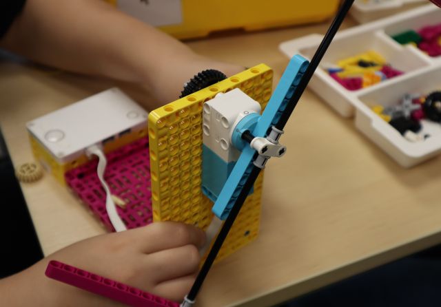 Na zdjęciu znajduje się zbudowany z klocków LEGO wiatrak, po jego bokach chłopiec trzyma ręce, na stoliku obok, wiatraka znajduje się plastikowe pudełko z klockami.
