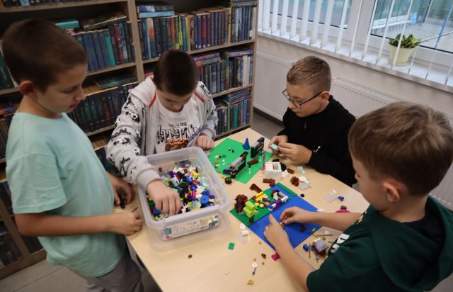 Na zdjęciu znajduje się 4 chłopców przy stoliku, przy którym wspólnie układają klocki LEGO, które znajdują się w plastikowym pudełku. W tle znajdują się półki z książkami i okna.