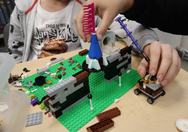 Na zdjęciu znajduje się mur zbudowany z klocków LEGO na zielonej podstawce, praca stoi na stole, a w tle siedzi chłopiec i dodaje kolejne elementy.