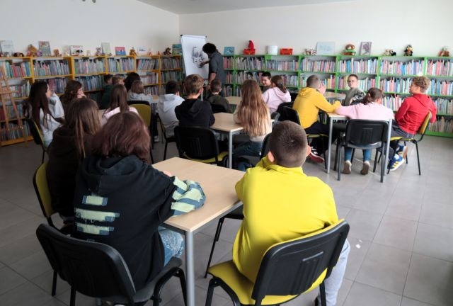 Na zdjęciu znajduje się 20 uczniów przy 6 stolikach, przed nimi stoi mężczyzna i rysuje na fliparcie. W tle znajdują się półki z książkami.