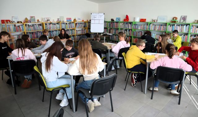 Na zdjęciu znajduje się 20 uczniów przy 5 stolikach i rysują na kartkach. W tle znajduje się flipart oraz półki z książkami.