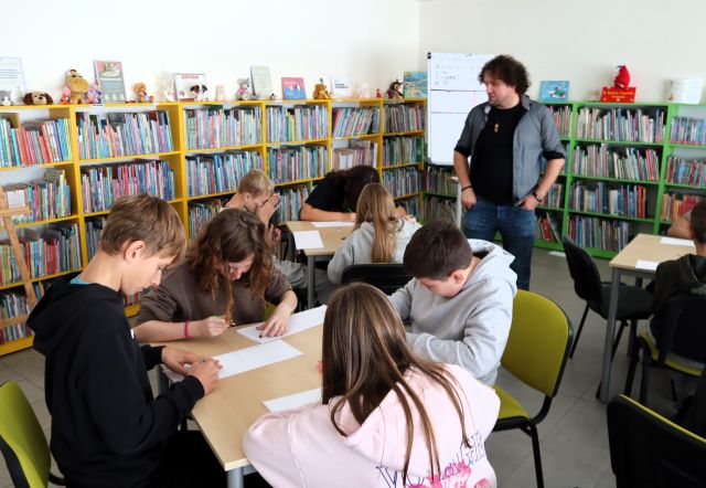 Na zdjęciu znajdują się dwa stoliki, przy których siedzą uczniowie i tworzą swój komiks na kartkach. Przy jednym stoliku stoi mężczyzna, a za nim znajduje się flipart oraz półki z książkami.