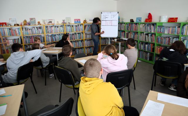 Na zdjęciu znajdują się uczniowie przy stolikach i patrzą na mężczyznę, który rysuje na fliparcie. W tle znajdują się półki z książkami.