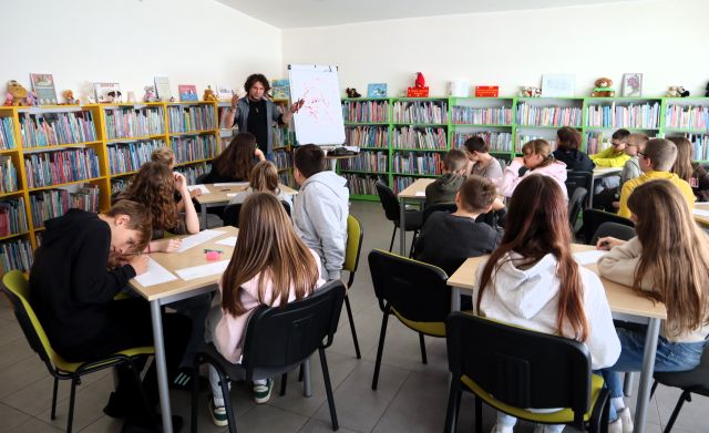 Na zdjęciu znajduje się 5 stolików, przy których siedzą uczniowie, przed nimi stoi mężczyzna i gestykuluje, po prawej stronie stoi flipart z namalowanym portretem. W tle znajdują się półki z książkami.