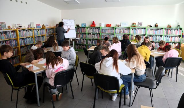 Na zdjęciu znajduje się 5 stolików, przy których siedzi 20 uczniów, część rysuje, część patrzy na mężczyznę przed nimi, który rysuje na fliparcie. W tle znajdują się półki z książkami.